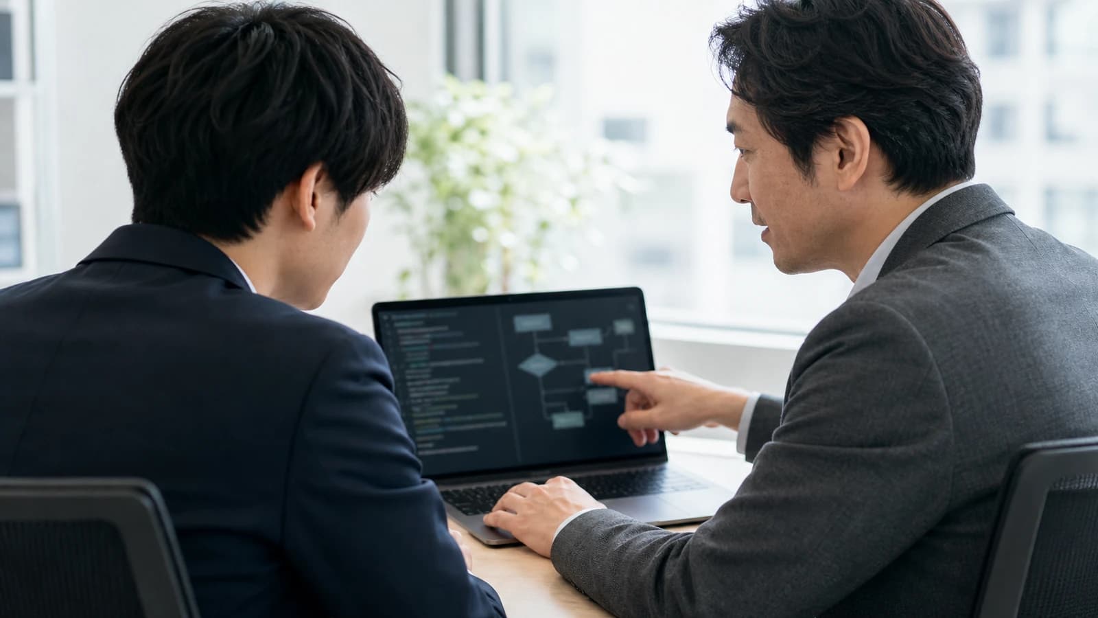 先輩社員がノートPCの画面を指しながら若手社員に説明している後ろ姿